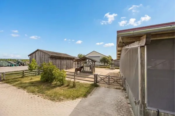Außenaufnahme eines Paddock-Bereichs auf einem Hofgut. Im umzäunten Sandbereich steht eine überdachte Holz-Heuraufe, bei der sich ein Pferd aufhält. Der Hof ist von rustikalen Holzgebäuden, Wiesen und Bäumen umgeben unter einem sonnigen, leicht bewölkten Himmel.