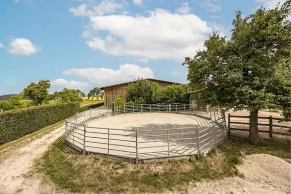 Außenaufnahme eines kreisrunden Reitplatzes (Roundpen) mit hellem Sandboden und einer Einfassung aus grauen Metall-Panels. Das Trainingsareal befindet sich auf einem ländlichen Anwesen vor einem hölzernen Stallgebäude, umgeben von Bäumen und einer grünen Hecke unter leicht bewölktem blauen Himmel.