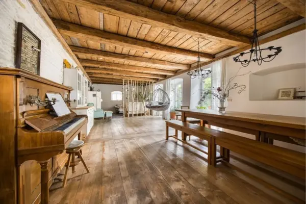 Lichtdurchfluteter, weitläufiger Wohnraum mit historischem Charme, geprägt von einer massiven Holzbalkendecke und warmem Dielenboden. Links im Vordergrund steht ein klassisches Holzklavier mit Hocker, rechts eine hölzerne Essgruppe mit Bänken. Im hinteren Bereich ergänzen ein moderner Hängesessel und ein Raumteiler aus Birkenstämmen die gemütliche Einrichtung.
