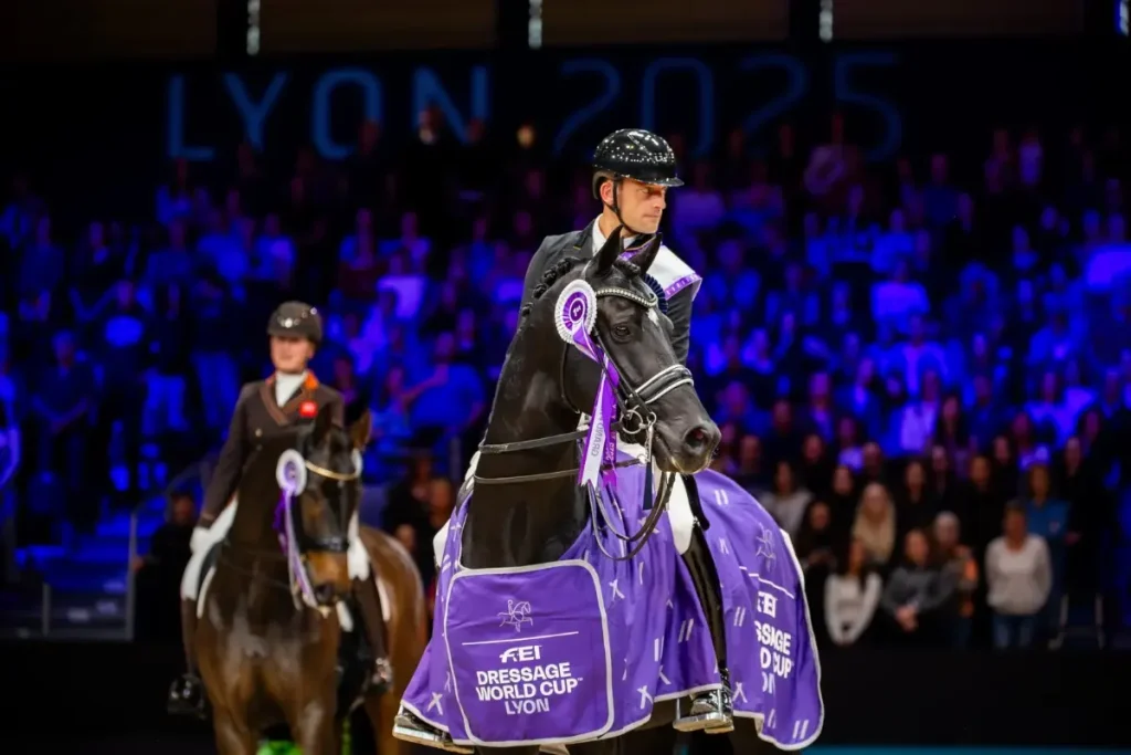 Dressurreiter Justin Verboomen auf dem eleganten Rappen Zonik Plus bei der Siegerehrung des FEI Dressage World Cup 2025 in Lyon. Das Pferd trägt eine violette Siegerdecke sowie eine Siegerschleife an der Trense; im Hintergrund reitet ein weiteres platziertes Paar vor vollbesetzten, bläulich beleuchteten Zuschauertribünen.