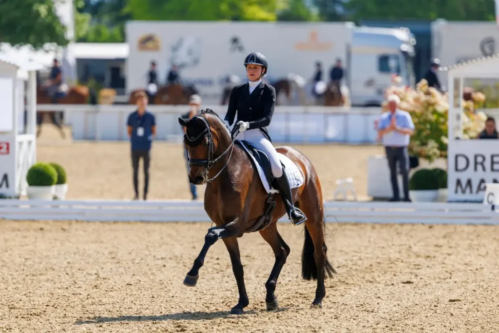 Ein Foto, das die U25-Dressurreiterin Katharina Schuster auf einem dunkelbraunen Pferd während einer Vorführung auf einem Sand-Dressurplatz zeigt. Die Reiterin trägt formelle Dressurkleidung, einschließlich eines schwarzen Fracks und eines Reithelms, und konzentriert sich auf die Übung. Das Pferd ist in einer fortgeschrittenen Dressurbewegung, wobei ein Bein angehoben ist. Der Hintergrund ist ein Wettbewerbsumfeld mit Zuschauern, Richterhäuschen und Werbebannern.