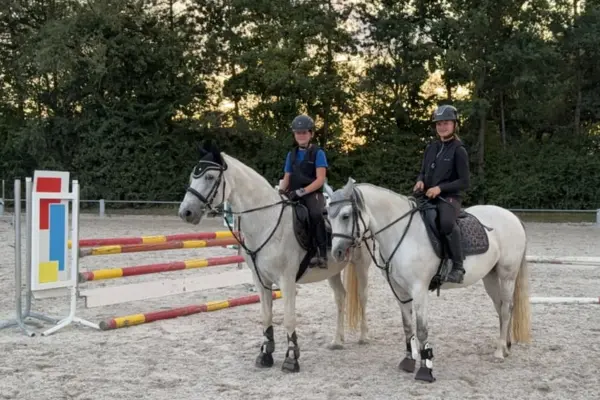 Ein Nahaufnahme-Foto von zwei jungen Reiterinnen, die nebeneinander auf zwei Schimmelpferden (weißen Pferden) auf einem Sandreitplatz stehen. Beide Reiterinnen tragen Helme und Sicherheitswesten und schauen lächelnd in die Kamera. Die Pferde haben Zaumzeug, Fliegenhauben und Beinschutz. Im Hintergrund links steht ein bunter Hindernisständer mit Springstangen vor einer Hecke und Bäumen.