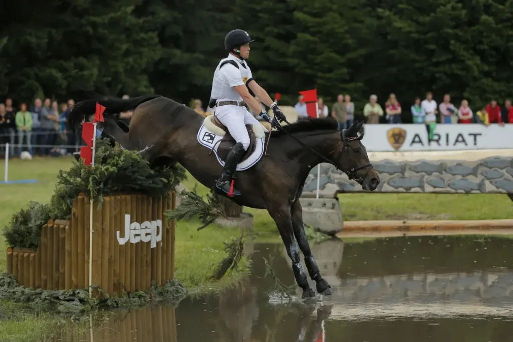 Der Vielseitigkeitsreiter Michael Jung springt mit einem dunkelbraunen Pferd über ein massives Holzhindernis mit Tannengrün und dem weißen „Jeep“-Schriftzug. Das Pferd landet mit den Vorderbeinen in einem Wasserkomplex. Im Hintergrund stehen Zuschauer hinter einer Absperrung, und eine Werbebande mit der Aufschrift „ARIAT“ ist vor einer dichten Baumreihe zu erkennen.