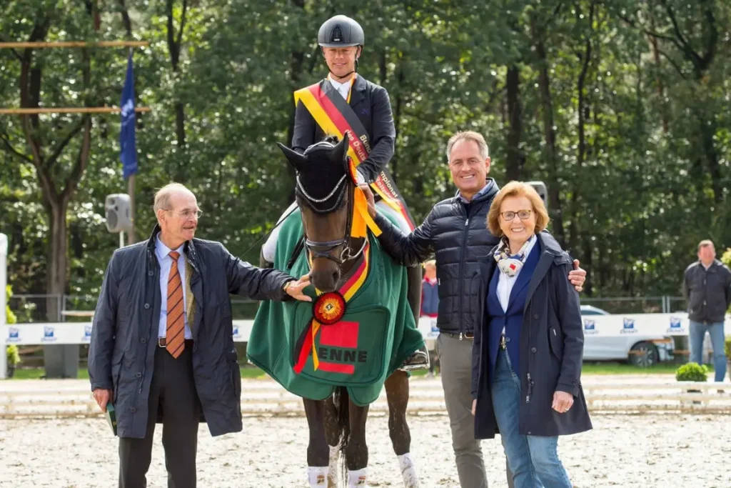 Siegerehrung auf einem Sand-Reitplatz. Im Zentrum steht das braune Pferd, Casey OLD, mit einer grünen Siegerdecke, die eine rot-gelbe Rosette und den Schriftzug 'ENNE' trägt. Hermann Gerdes, der Reiter, sitzt im schwarzen Dressur-Outfit mit einer schwarz-rot-goldenen Schärpe über der Schulter auf dem Pferd. Flankiert wird die Gruppe von drei Personen: links Paul Wendeln im dunklen Mantel, rechts Peter Wendeln in einer Daunenjacke und Hilde Wendeln im blauen Mantel und bunten Schal, die alle lächeln. Der Hintergrund zeigt grüne Bäume und Flaggen.
