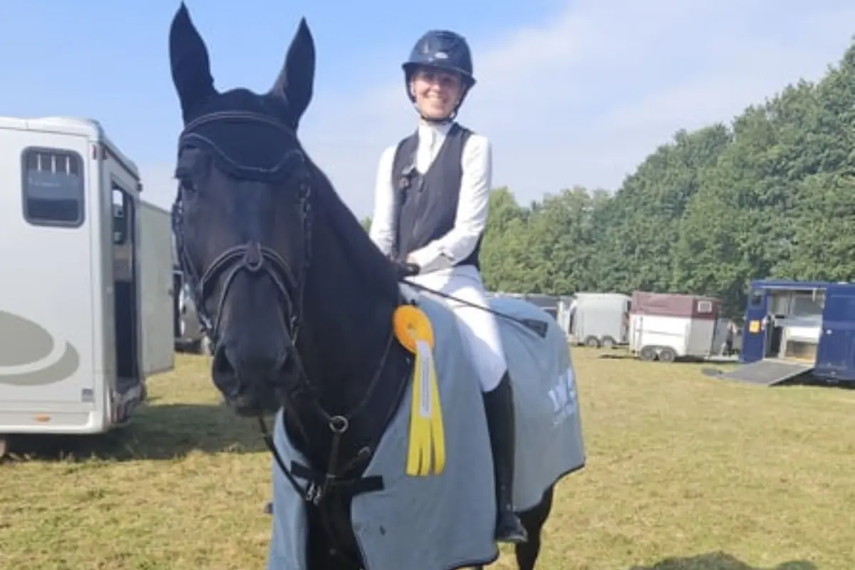 S*-Sieger · CSI1* · Amateurgeeignet: Reiterin mit Pferd auf Turniergelände, Pferd hat Siegerschleife an der Decke, beide schauen zur Kamera