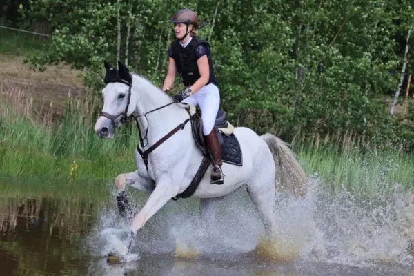 Dynamische Aufnahme des Schimmels, wie er kraftvoll durch ein Wasserhindernis galoppiert. Das Wasser spritzt hoch auf, während das Pferd mit hoher Konzentration den Weg durch den Teich sucht. Die Reiterin sitzt ausbalanciert im leichten Sitz.
