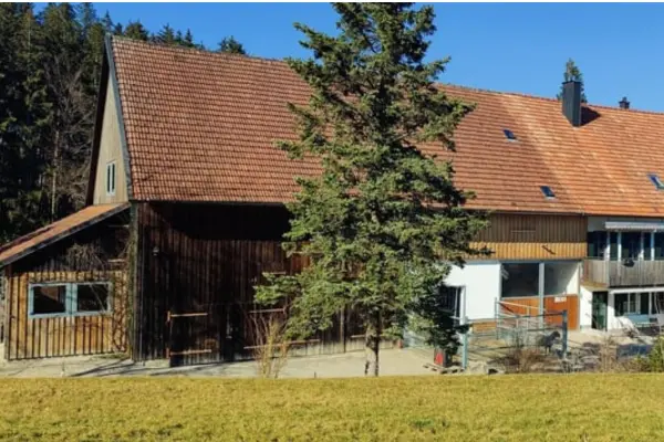 Traditionelles Stall- und Wohngebäude eines Reitanwesens mit rotem Ziegeldach, dunkler Holzfassade und Paddockbereich vor einer Waldkulisse.