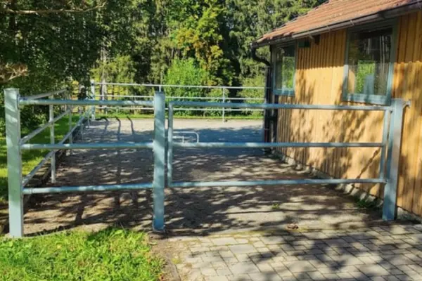 Ein befestigter Paddockauslauf mit silbernem Metallzaun direkt an einem Gebäude mit hellbrauner Holzverkleidung.
