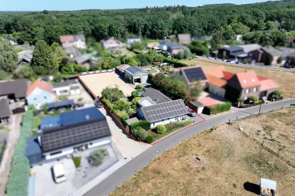 Eine Schrägluftaufnahme mit Tilt-Shift-Effekt zeigt ein ländliches Anwesen in Deutschland. Das Grundstück ist mit einer roten Linie umrandet und umfasst ein Wohnhaus mit Photovoltaikanlage, einen großen Sandreitplatz, ein Nebengebäude (Stall/Halle) mit Photovoltaikanlage, Gärten und Bäume. Die Umgebung ist unscharf dargestellt, was den Fokus auf das hervorgehobene Grundstück lenkt.