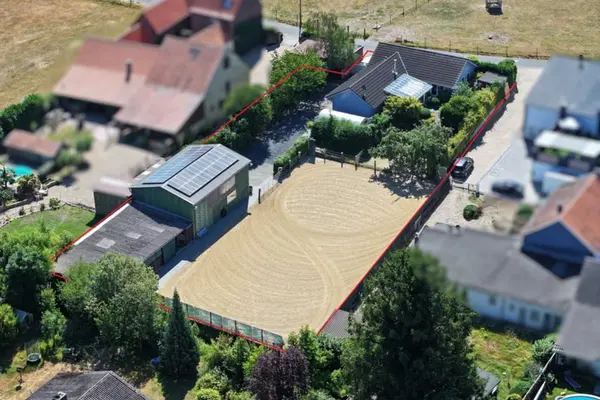 Eine Schrägluftaufnahme mit Tilt-Shift-Effekt zeigt ein ländliches Anwesen in Deutschland. Das Grundstück ist mit einer roten Linie umrandet und umfasst ein Wohnhaus mit Photovoltaikanlage, einen großen Sandreitplatz, ein Nebengebäude (Stall/Halle) mit Photovoltaikanlage, Gärten und Bäume. Die Umgebung ist unscharf dargestellt, was den Fokus auf das hervorgehobene Grundstück lenkt.