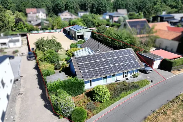 Eine Schrägluftaufnahme mit Tilt-Shift-Effekt zeigt ein ländliches Anwesen in Deutschland. Das Grundstück ist mit einer roten Linie umrandet und umfasst ein Wohnhaus mit Photovoltaikanlage, einen großen Sandreitplatz, ein Nebengebäude (Stall/Halle) mit Photovoltaikanlage, Gärten und Bäume. Die Umgebung ist unscharf dargestellt, was den Fokus auf das hervorgehobene Grundstück lenkt.