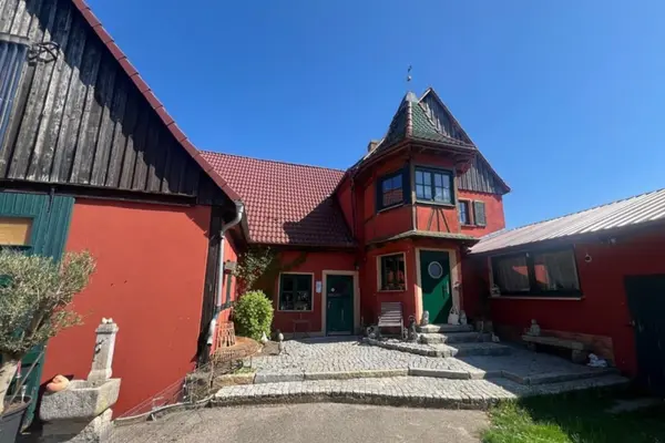 Außenansicht eines idyllischen, rot verputzten Fachwerkhauses mit dunkelgrünen Fenstrahmen und Türen. Das Gebäude hat ein rotes Ziegeldach und rechts einen angebauten Turm mit Erkerfenstern. Vor dem Eingang befindet sich ein kleiner, mit Kopfsteinpflaster ausgelegter Hof mit Pflanzen, einer Gartenbank und Deko-Elementen bei strahlendem Sonnenschein.