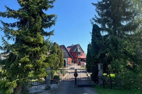 Ansicht in einen Hof eines idyllischen, rot verputzten Fachwerkhauses mit dunkelgrünen Fenstrahmen und Türen. Das Gebäude hat ein rotes Ziegeldach und rechts einen angebauten Turm mit Erkerfenstern. Vor dem Eingang befindet sich ein kleiner, mit Kopfsteinpflaster ausgelegter Hof mit Pflanzen, einer Gartenbank und Deko-Elementen bei strahlendem Sonnenschein.