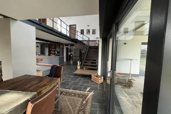 Innenansicht eines modernen, offenen Wohnbereichs im Loft-Stil mit dunklem Schieferfliesenboden. Im Vordergrund steht ein hölzerner Esstisch mit braunen Lederstühlen auf einem Orientteppich. Eine markante schwarze Stahltreppe mit Geländer führt auf eine offene Galerie. Im Hintergrund ist eine minimalistische, anthrazitfarbene Küche mit Kochinsel zu sehen, rechts eine große Glasschiebetür nach draußen.
