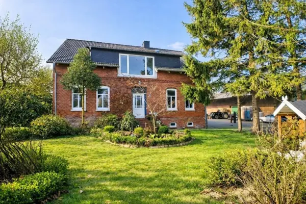 Die Frontansicht eines charmanten, traditionellen Backstein-Wohnhauses mit rotem Klinker und einem gepflegten grünen Vorgarten samt angelegtem Beet. Das Haus verfügt über weiße Fensterrahmen, eine weiße Eingangstür und ein ausgebautes Dachgeschoss. Auf der rechten Seite führt eine gepflasterte Zufahrt vorbei an hohen Nadelbäumen zum Hofbereich, wo Ställe und ein Traktor zu sehen sind.