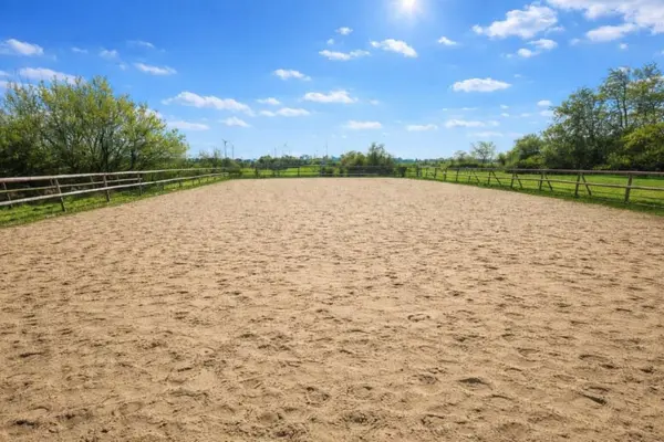 Ein großzügiger Außenreitplatz mit Sandboden an einem sonnigen Tag. Der Platz ist komplett mit einem klassischen Holz-Koppelzaun eingefasst. Im Hintergrund sieht man grüne Wiesen, Bäume und vereinzelte Windkraftanlagen am Horizont unter einem strahlend blauen Himmel mit lockeren Schönwetterwolken.