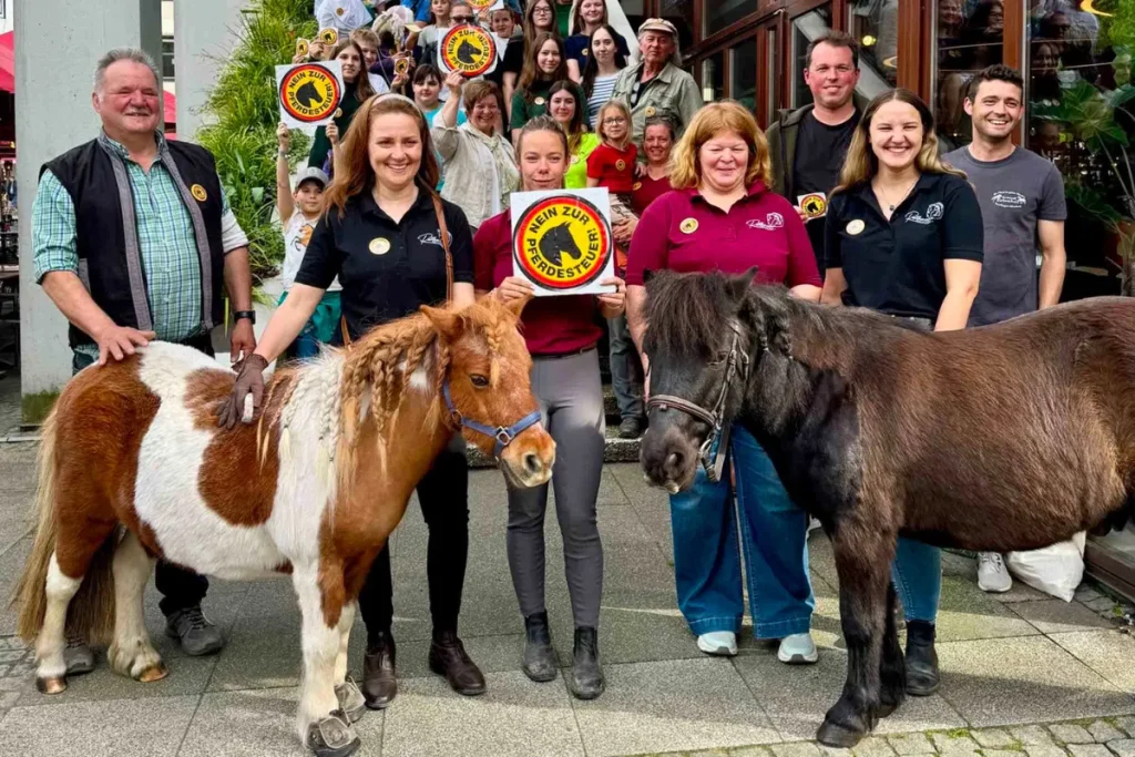 Eine Gruppe von Menschen protestiert in Reutlingen gegen die Pferdesteuer. Im Vordergrund stehen zwei kleine Ponys – links ein braun-weiß geschecktes mit geflochtener Mähne, rechts ein dunkelbraunes. Eine junge Frau in der Mitte hält ein quadratisches Schild mit der Aufschrift „NEIN ZUR PFERDESTEUER!“ und einem durchgestrichenen Pferdekopf-Symbol in die Kamera. Im Hintergrund stehen weitere lächelnde Teilnehmende auf einer Treppe, von denen einige kleinere Versionen des Protest-Logos hochhalten.
