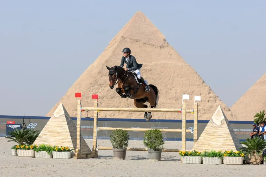 Springreiter Pieter Devos überwindet im Springsitz auf dem braunen Pferd Casual DV Z ein thematisch gestaltetes Hindernis vor der monumentalen Kulisse der Pyramiden von Gizeh. Foto: LGCT/Stefano Grasso