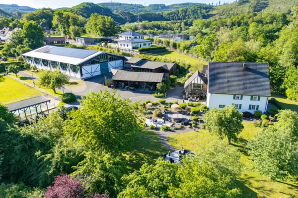 Luftaufnahme aus der Vogelperspektive einer weitläufigen, idyllischen Reitanlage in Plettenberg (Sauerland). Auf der rechten Seite befindet sich ein großes, weißes Wohnhaus mit dunklem Giebeldach und grünem Fachwerk-Anbau, umgeben von einem weitläufigen Garten mit Terrasse, dichtem Baumbestand und Rasenflächen. Im Zentrum und auf der linken Seite erstrecken sich die funktionalen Reitsportgebäude, darunter eine große, moderne Reithalle mit PV-Anlage auf dem Dach und markanter Fachwerkfassade, diverse Stallungen, Offenunterstände sowie gepflasterte Hof- und Zufahrtswege. Eingebettet ist das gesamte Anwesen in eine hügelige, waldreiche Landschaft mit grünen Wiesen und bewaldeten Hängen im Hintergrund unter sonnigem, blauem Himmel.