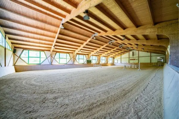 Innenansicht der Reithalle auf dem Anwesen in Plettenberg mit sandigem Reitboden. Die große Halle ist von einer hohen Dachkonstruktion aus Naturholz mit massiven Leimholzbindern und Deckenleuchten überspannt. Auf der linken Seite befindet sich eine weiß verputzte Wand mit großen, sechseckigen Fenstern, die grüne Rahmen haben und natürliches Tageslicht hereinlassen. Die verkleideten Banden umschließen die sandige Reitfläche, auf der im hinteren Bereich mehrere Springhindernisse und Cavalettis aufgebaut sind. Am Ende der Halle führt eine Tür in den angrenzenden Stalltrakt.