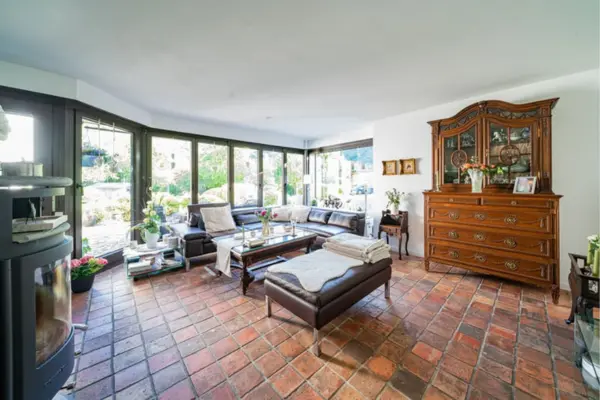 Heller, gemütlicher Wohnbereich im Wintergarten des Anwesens in Plettenberg mit rustikalem Terracotta-Fliesenboden und großzügiger Panorama-Glasfront zum grünen Garten. Im linken Vordergrund befindet sich ein moderner schwarzer Kaminofen. Der Raum ist eingerichtet mit einer dunklen Leder-Eckcouch, einem passenden Leder-Hocker sowie einem aufwendig geschnitzten, historischen Holzschrank an der rechten Wand.