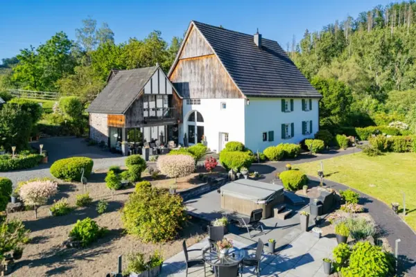 Luftaufnahme eines idyllischen Anwesens in Plettenberg (Sauerland) mit einem großen historischen Wohngebäude mit weißer Fassade und hohem Giebeldach, an das sich ein rustikaler Fachwerk- und Holzanbau anschließt. Im gepflegten, parkähnlichen Garten im Vordergrund befinden sich eine gepflasterte Terrasse mit Sitzgelegenheiten, ein im Boden eingelassener Whirlpool, formvollendete Sträucher und Büsche sowie weite Rasenflächen und dichter Baumbestand im Hintergrund bei sonnigem Tageslicht.