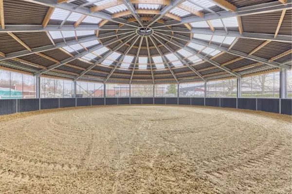 Innenansicht einer kreisförmigen, überdachten Reithalle (Longierhalle). Der helle Sandboden zeigt deutliche Traktorspuren. Eine niedrige schwarze Bande und eine Panoramaufensterfront umschließen die Bahn. Das Dach ist eine radial-symmetrische Holz- und Metallkonstruktion mit integrierten Lichtbändern. Künstliche Leuchten sind ebenfalls vorhanden.