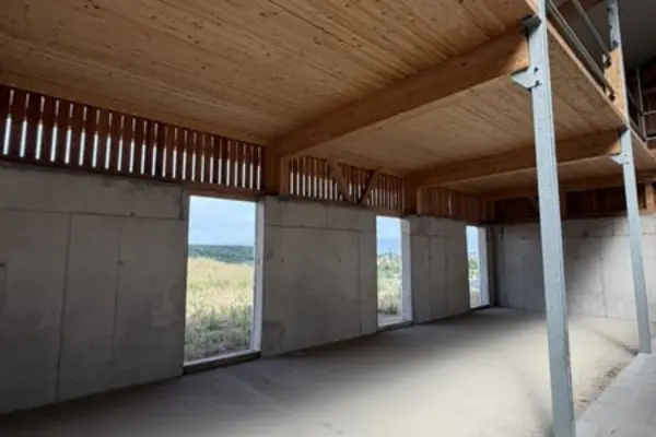 Blick von innen in einen hellen, noch unmöblierten Stallneubau. Die Außenwände bestehen im unteren Bereich aus grauem Sichtbeton mit mehreren hohen Fensterausschnitten, die den Blick auf eine Wiesenlandschaft freigeben. Im oberen Bereich sorgen vertikale Holzlamellen für Durchlüftung unter einer massiven Holzbalkendecke.