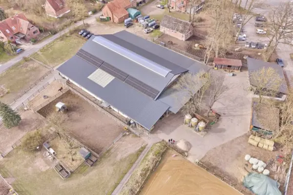 Vogelperspektive einer Reitanlage mit einem großen zentralen Hallen- und Stallgebäude, dessen dunkles Dach mit Lichtbändern und Solarmodulen ausgestattet ist. Rund um den Gebäudekomplex befinden sich eingezäunte Sandpaddocks, Futtersilos, gelagerte Heuballen sowie ein Teil eines Reitplatzes, eingebettet in eine ländliche Umgebung mit angrenzenden Wohnhäusern und Straßen.