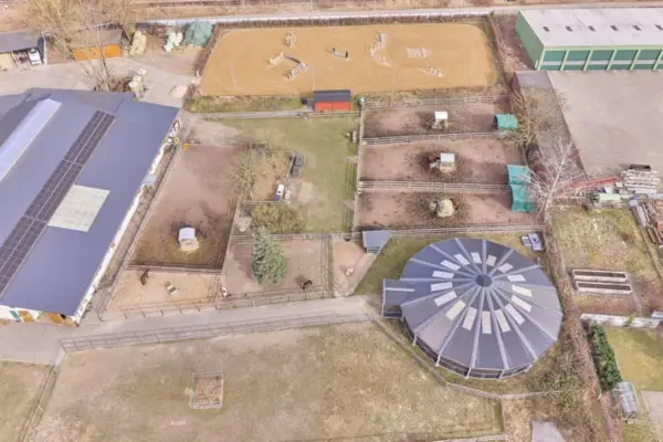 Luftaufnahme einer Reitanlage aus der Vogelperspektive mit einer großen Reithalle samt Solarmodulen auf der linken Seite, einer runden, überdachten Longierhalle unten rechts, einem Sandreitplatz mit Springhindernissen oben sowie mehreren eingezäunten Paddocks in der Mitte.