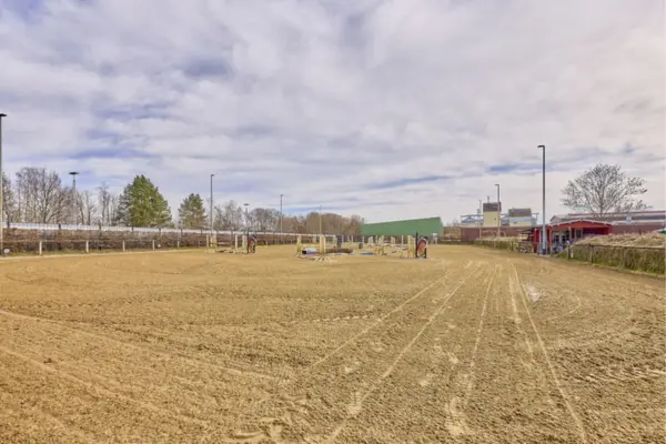 Weitläufiger Reitplatz mit ebenem Sandboden und verschiedenen aufgebauten Springhindernissen in der Mitte. Der Platz ist von Umzäunungen sowie Flutlichtmasten umgeben; im Hintergrund befinden sich ein kleines rotes Gebäude, Bäume und ein leicht bewölkter Himmel.