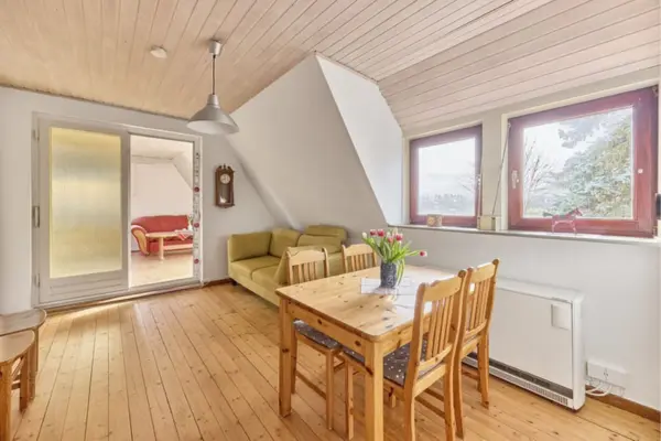 Hell eingerichtetes Zimmer einer Dachgeschosswohnung mit Holzdielenboden und heller Holzdeckenverkleidung. Im Vordergrund steht ein Holztisch mit Stühlen und einer Blumenvase, dahinter ein grünlich-gelbes Sofa unter einer Dachschräge. Rechts befinden sich zwei Fenster mit braunem Rahmen und ein Heizkörper, während links eine Glasschiebetür den Blick in einen angrenzenden Raum mit rotem Sofa freigibt.