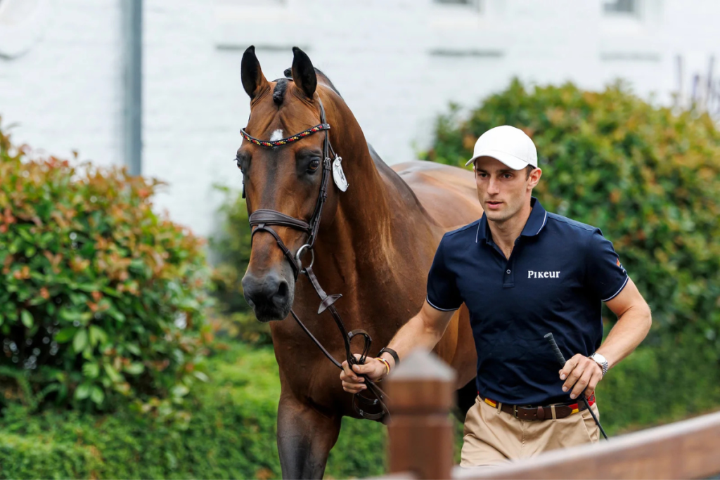 Der Springreiter Richard Vogel führt ein braunes Pferd an den Zügeln. Er trägt ein dunkelblaues Polo-Shirt der Marke Pikeur, eine weiße Basecap und beige Hosen und blickt konzentriert nach vorne. Das edle Pferd läuft aufmerksam neben ihm, trägt eine Trensenzaumzeug mit verziertem Stirnriemen und blickt in die Kamera. Im Hintergrund befinden sich grüne Büsche und die weiße Fassade eines Gebäudes.