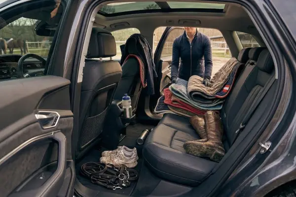 Blick in den Innenraum eines modernen Autos, das als mobiler Sattelschrank dient. Auf der Rückbank stapeln sich mehrere bunte Schabracken und ein Paar getragene Leder-Reitstiefel. Im Fußraum stehen schmutzige Turnschuhe neben einer abgelegten Trense. Eine Reiterin in dunkler Funktionsjacke ist im Anschnitt zu sehen, wie sie gerade Ordnung schafft. Im Hintergrund ist ein Paddock mit Pferden erkennbar. Das Bild vermittelt den authentischen, leicht chaotischen Stallalltag.