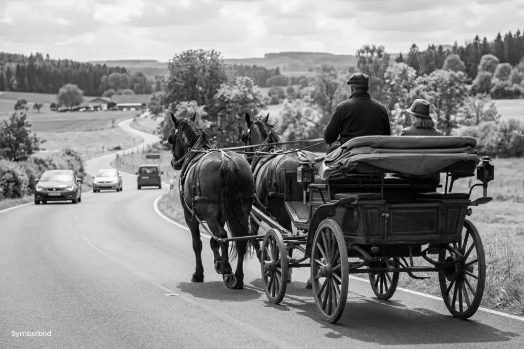 Eine Rückansicht eines hölzernen Kutschgespanns mit zwei braunen Pferden, das auf der rechten Seite einer asphaltierten Landstraße fährt. Auf dem Bock sitzen ein Mann mit Schirmmütze und eine Frau mit Hut. Die Straße windet sich durch eine grüne, hügelige Landschaft mit Feldern und Bäumen. In der Ferne kommen dem Gespann auf der linken Fahrbahnseite mehrere moderne Autos entgegen.