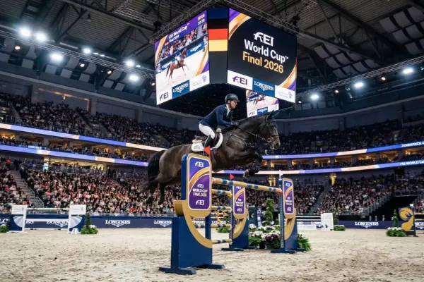 Eine dynamische Sportfotografie zeigt einen Springreiter und sein braunes Pferd im Moment des Sprungs über ein blau-goldenes FEI-Hindernis. Das Paar befindet sich mitten in der Luft über den Stangen. Der Wettbewerb findet in einer riesigen, voll besetzten Indoor-Arena statt. Über der Reitbahn hängt ein großer Videowürfel, der das Logo des "FEI World Cup™ Final 2026" sowie die deutsche Flagge zeigt. Die Zuschauerränge im Hintergrund sind dicht gefüllt, und die Arena wird durch helles Flutlicht beleuchtet. Der Boden ist mit hellem Reitsand bedeckt.