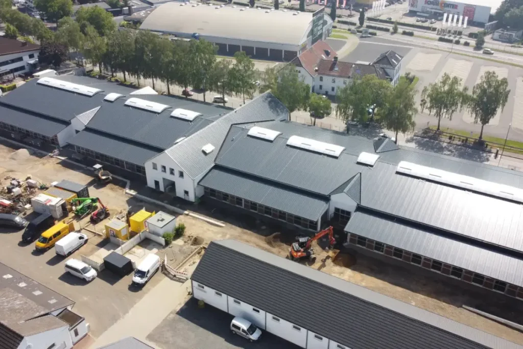 Luftaufnahme einer Großbaustelle für moderne Stallneubauten der Reitsport-WM in Aachen. Zu sehen sind weitläufige Gebäude mit grauen Dächern und Oberlichtern, umgeben von Baggern, Lieferwagen und Baumaterialien in einem städtischen Gewerbegebiet mit Parkplätzen und Bäumen.