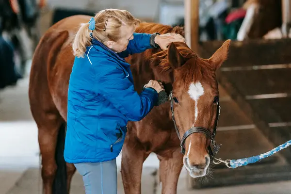 Eine ältere Frau mit blondem Pferdeschwanz in einer blauen Jacke striegelt liebevoll die Mähne eines braunen Pferdes mit einer weißen Blesse. Das Pferd steht in einem Stallgang und schaut zur Kamera. Sie massiert den Hals und benutzt eine Bürste.