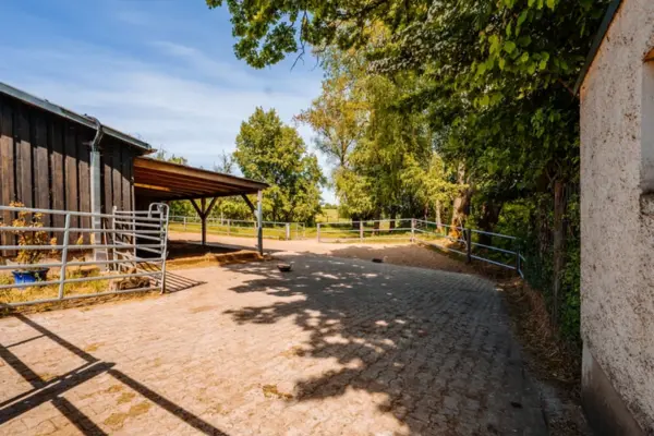 Gepflasterter Außenbereich einer Reitanlage mit dunklem Holzstall, überdachtem Unterstand und Koppelzäunen unter sonnigem Himmel.