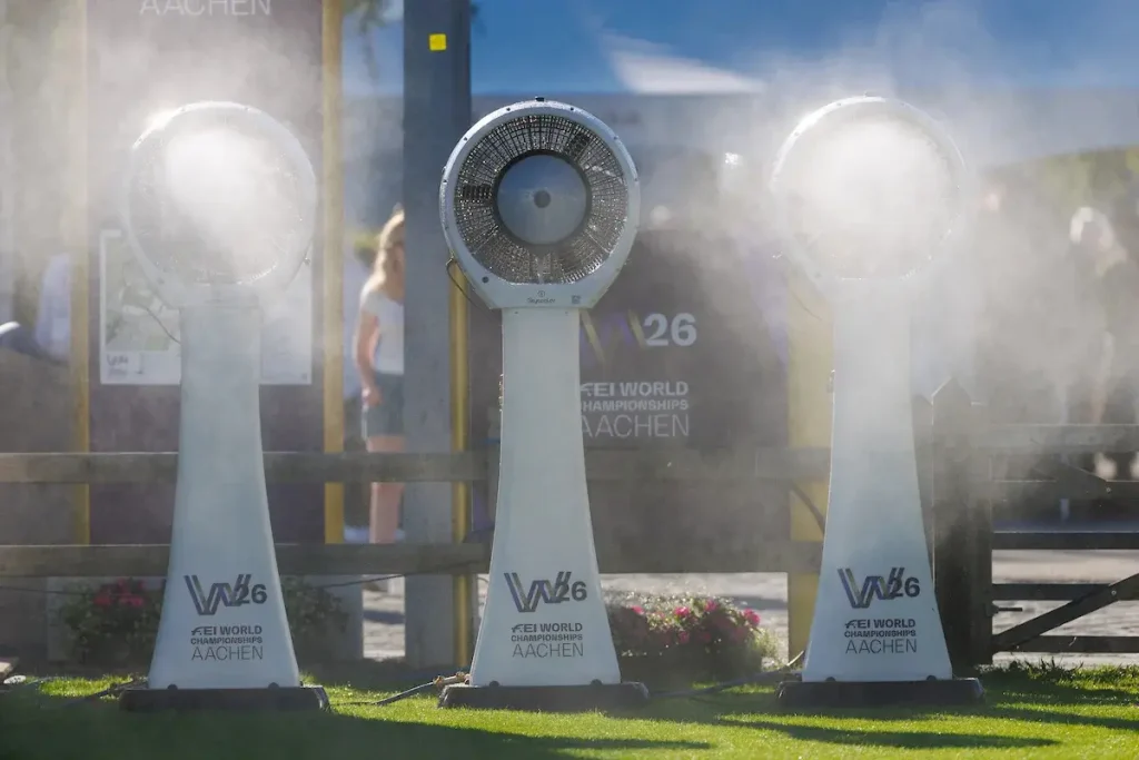 Drei große weiße Standventilatoren mit dem Logo der FEI World Championships Aachen 2026, die auf einer Wiese feinen Kühlnebel versprühen.