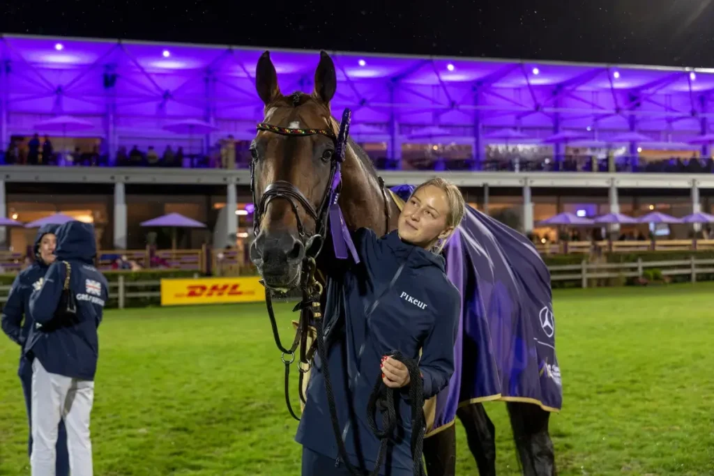 Pflegerin Felicia Wallin steht lächelnd neben dem braunen Hengst United Touch S auf dem Rasenplatz; das Pferd trägt Zaumzeug mit lila Schleife und eine Abschwitzdecke vor einer violett beleuchteten Stadiontribüne bei Nacht.