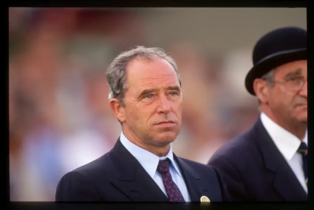 Ein Farbfoto aus dem Jahr 1992, das Uwe Mechlem zeigt. Er ist ein Mann mittleren Alters mit ernstem Gesichtsausdruck, der nach rechts blickt. Er trägt einen dunklen Anzug, ein hellblaues Hemd und eine rote Krawatte mit einem goldfarbenen Hufeisenmuster. Am Revers ist ein Abzeichen angebracht. Rechts von ihm ist teilweise ein weiterer Mann mit Brille und einem schwarzen Melone-Hut zu sehen. Der Hintergrund ist eine unscharfe Menschenmenge bei einer Freiluftveranstaltung, und das Foto hat einen dunklen Rand.