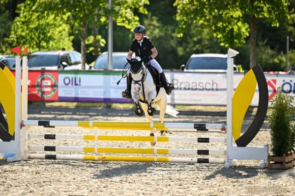 Eine junge Reiterin auf einem Schimmelpony springt bei einem Springturnier über ein weiß-gelb-schwarzes Hindernis. Das Pony ist mitten im Sprung über der obersten Stange, alle vier Beine sind angezogen. Die Reiterin trägt einen schwarzen Helm, eine schwarze Jacke, weiße Hosen und Stiefel. Im Hintergrund sind Werbebanner, parkende Autos und Bäume auf einem Sandplatz zu sehen.